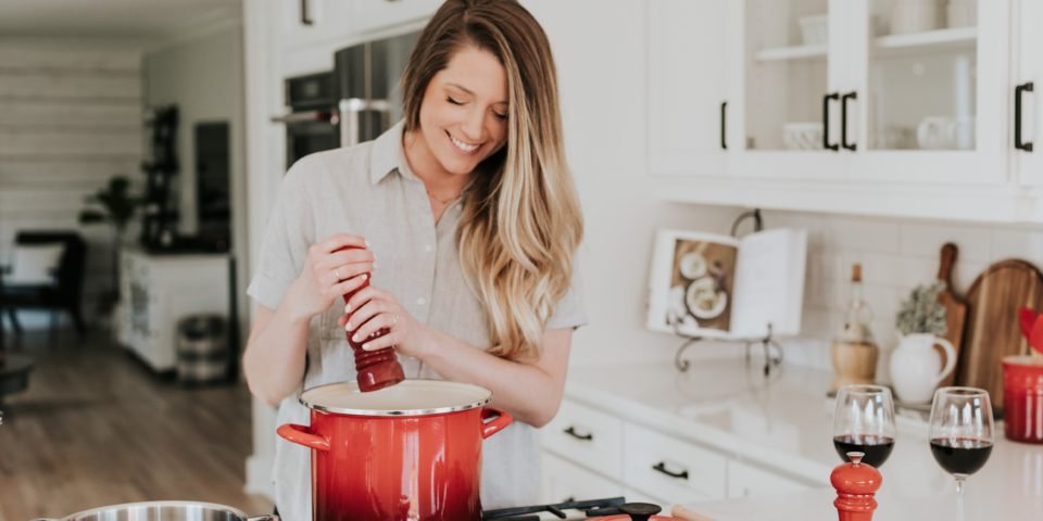 woman holding pepper grinder above a red pot in a kitchen